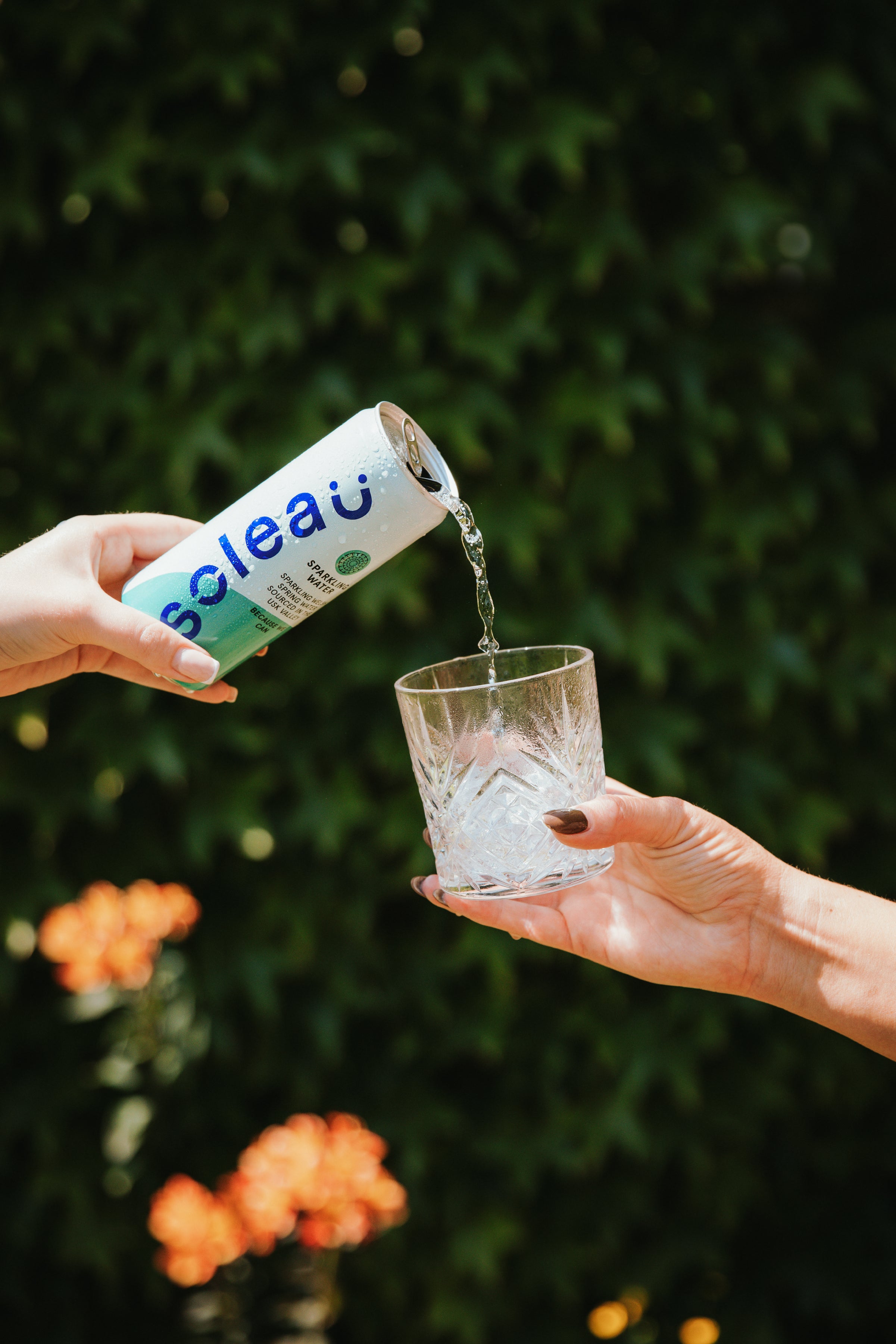 Person pouring water from a 'Soleau' can into a glass against a blurred green background with flowers.